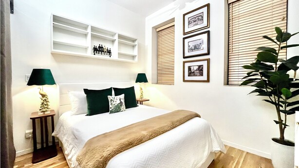 Une chambre à coucher avec un lit, des tables de chevet, une étagère, une fenêtre avec des stores et une plante en pot.