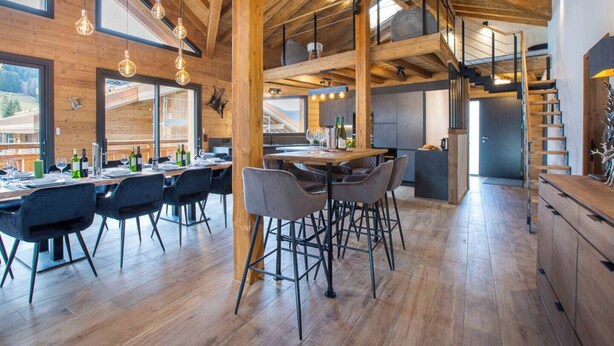 A spacious dining area with a high ceiling and exposed wooden beams, a kitchen area with modern appliances, and a staircase leading to an upper level. - Chalet Le Pre hosted by More Mountain Morzine | Luxury 6 Bed 6 Bath Chalet | Sleeps 14. Only 10 min walk into the heart of Morzine with huge outdoor Jacuzzi, large terrace & garden with stunning views of the slopes. Secluded position, yet central. (Morzine)