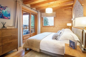 A cozy bedroom with a wooden ceiling, a large bed with white bedding, a nightstand with a lamp, and a window with curtains.