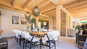A dining area with a wooden table, chairs with white fur throws, hanging wicker light fixtures, and a staircase leading to another room. - Chalet Verjus hosted by More Mountain, Morzine | Luxury 5 Bed 5 Bath Chalet | Sleeps 10. Luxury stand alone ski chalet in a very central location with Outdoor Jacuzzi & large terrace. All Mod Cons. Less than 5 mins walk to Ski Lifts. (Morzine)