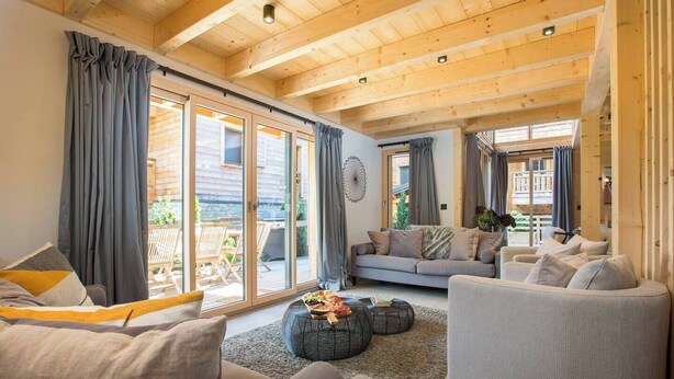 A spacious living room with a high wooden ceiling, large sliding glass doors leading to an outdoor area, and a cozy seating arrangement with sofas and armchairs. - Chalet Verjus hosted by More Mountain, Morzine | Luxury 5 Bed 5 Bath Chalet | Sleeps 10. Luxury stand alone ski chalet in a very central location with Outdoor Jacuzzi & large terrace. All Mod Cons. Less than 5 mins walk to Ski Lifts. (Morzine)