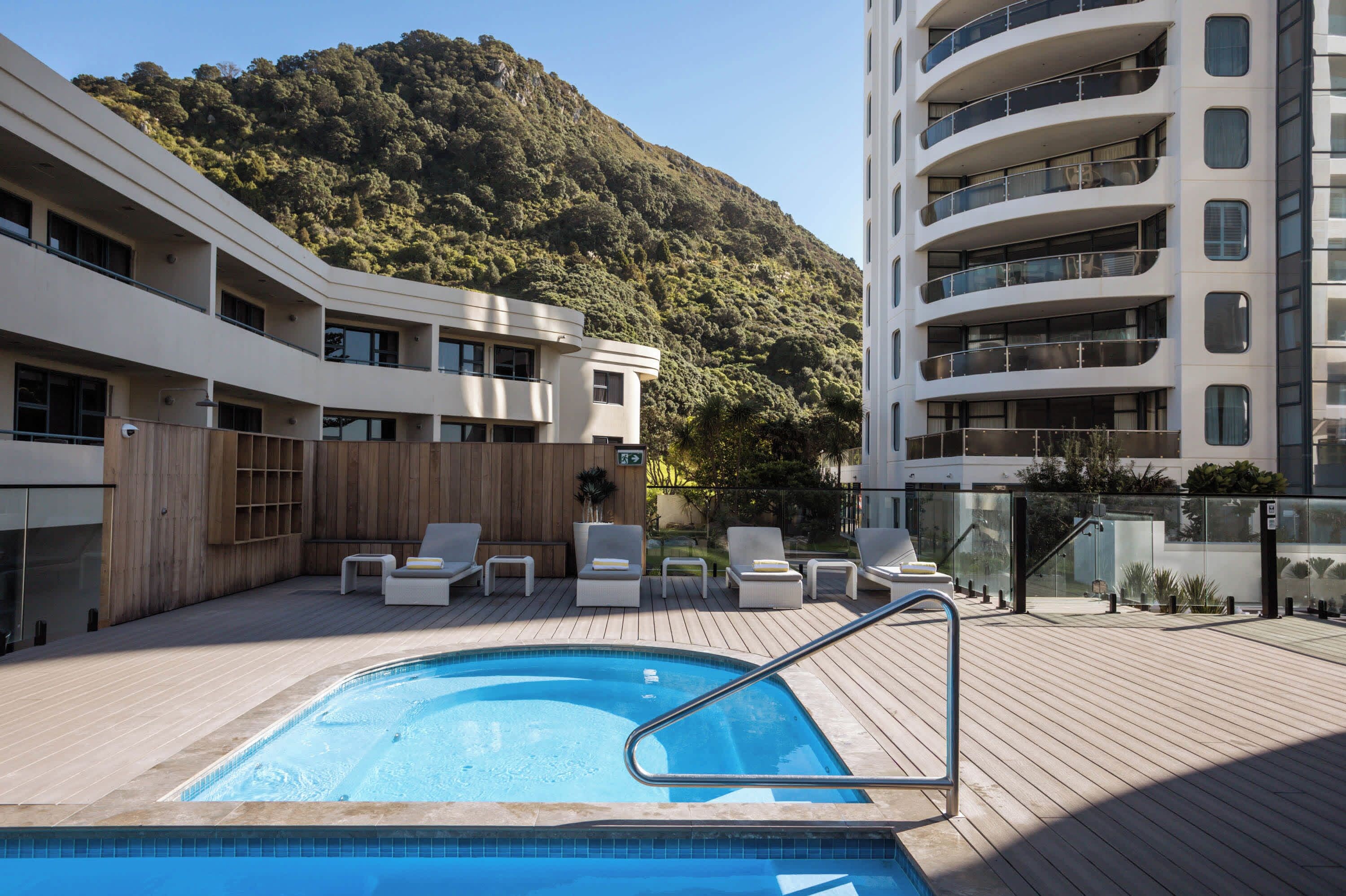 A rooftop pool with lounge chairs, a wooden deck, and a mountainous backdrop.