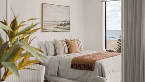 A bedroom with a large window, a bed with a beige blanket, and a painting on the wall.