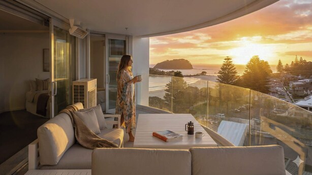 A modern living room with a glass balustrade, a sofa, a coffee table with a book and a coffee pot, and a person holding a cup.