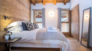 Un dormitorio con techo de madera, cama con edredón blanco y almohadas estampadas, mesita de noche con cafetera y tazas, y ventana con cortinas.