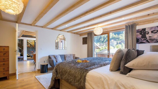 A bedroom with a large bed, a wooden ceiling, a view of the outdoors, and a dining area visible through an open door. - Apartment Grand Central hosted by More Mountain, Morzine | Luxury 3 Bed 3 Bath | Sleeps 6. Luxurious and Spacious duplex Penthouse apartment in the centre of Morzine, only 2 minutes walk on the flat to both the Pleney and Super Morzine Telecabines. (Morzine)