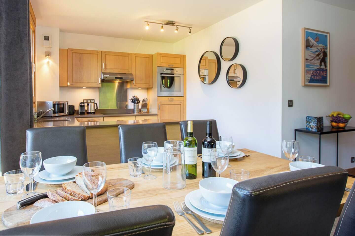 Uma sala de jantar moderna com uma mesa de madeira posta para seis pessoas, rodeada por cadeiras de couro. Uma cozinha com armários de madeira e eletrodomésticos de aço inoxidável é visível ao fundo.