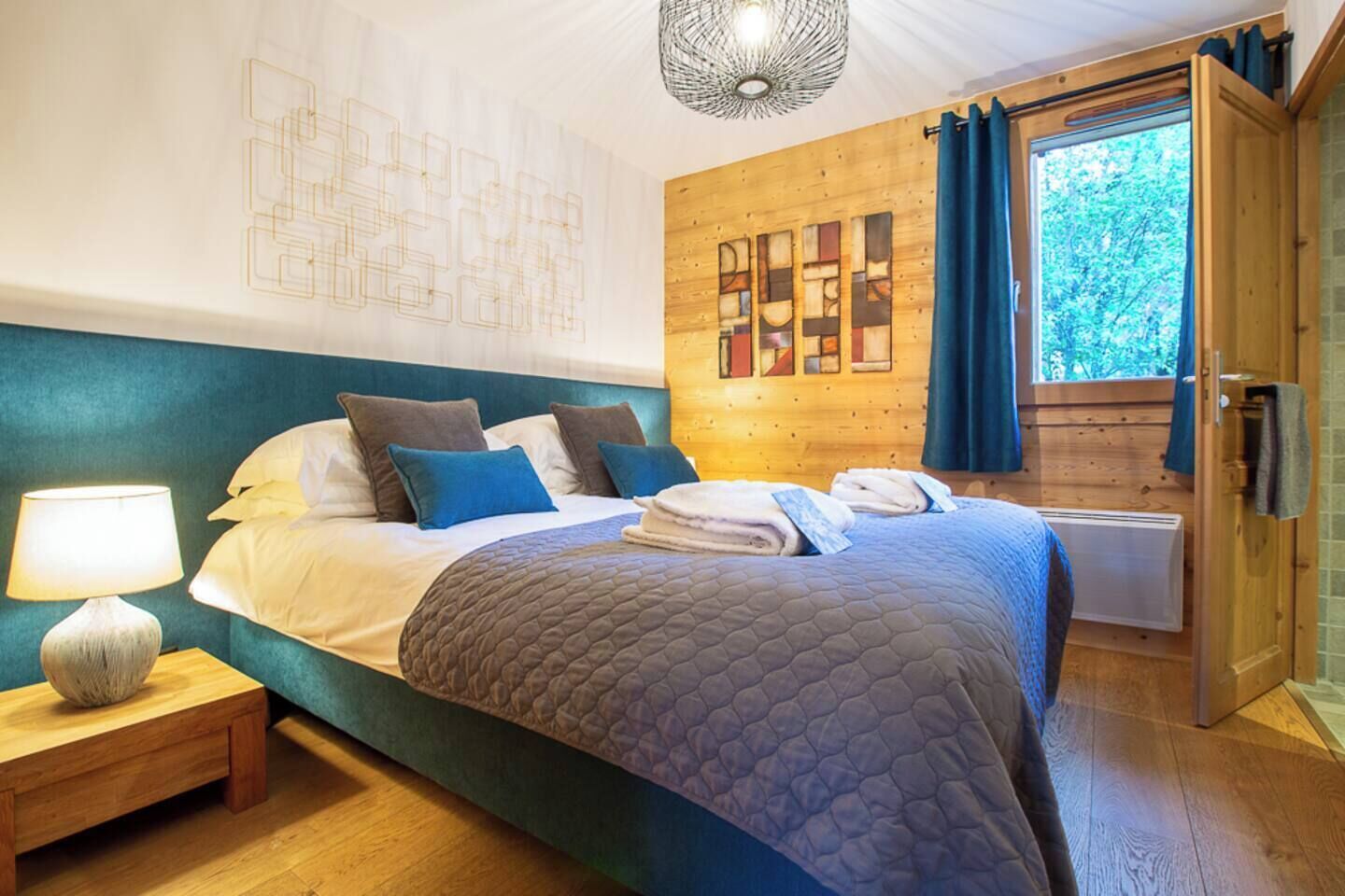 A bedroom with a large bed, a bedside table with a lamp, a wooden headboard, a wall with abstract art, and a window with curtains.