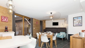 A modern dining area with a wooden table set for four, white chairs, and a ceiling fan. - Apartment La Poste hosted by More Mountain, Morzine | Large 3 Bed 2 Bath apartment | Sleeps 6 . Fantastic location with  large terrace in the centre of town. Family friendly with All Mod Cons. Only 5 mins walk to Ski Lifts and (Morzine)