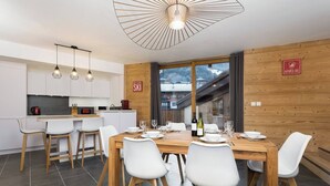 A modern kitchen with white cabinets, a wooden dining table set for four, and a view of a ski resort through large windows. - Apartment La Poste hosted by More Mountain, Morzine | Large 3 Bed 2 Bath apartment | Sleeps 6 . Fantastic location with  large terrace in the centre of town. Family friendly with All Mod Cons. Only 5 mins walk to Ski Lifts and (Morzine)