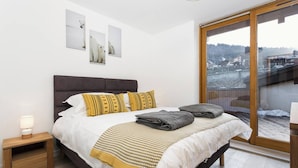 A modern bedroom with a large bed, a nightstand, a window with a view of a snowy landscape, and a ceiling light. - Apartment La Poste hosted by More Mountain, Morzine | Large 3 Bed 2 Bath apartment | Sleeps 6 . Fantastic location with  large terrace in the centre of town. Family friendly with All Mod Cons. Only 5 mins walk to Ski Lifts and (Morzine)