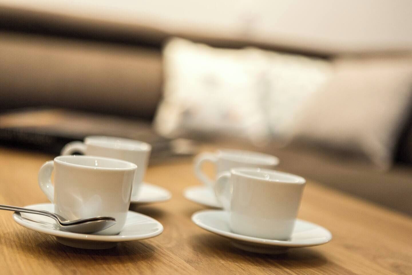 A series of white cups on saucers arranged on a wooden table, with a blurred background featuring a sofa and a pillow.