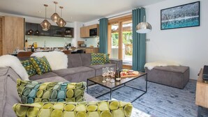 Una sala de estar moderna con un sofá gris, sillones estampados, una mesa de centro con una botella de vino y copas, y una cocina al fondo.