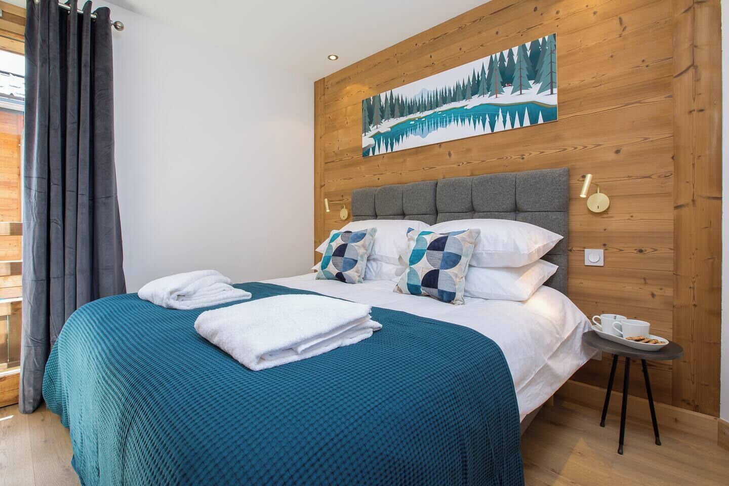 Un lit bien fait, recouvert d’une couvre-lit turquoise, avec des oreillers blancs et une tête de lit grise. Un tableau représentant un paysage enneigé est accroché au mur au-dessus du lit.