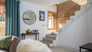 Una sala moderna con un sofá, un plaid blanco mullido, un espejo redondo y una pequeña mesa con frutas y una radio. Una escalera con barandillas de vidrio conduce a un altillo de madera.