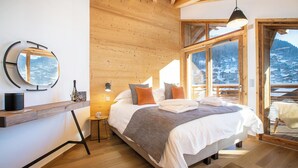 Ein modernes Schlafzimmer mit Holzboden, einem großen Bett mit Kissen, einem Nachttisch mit Lampe und Blick auf eine verschneite Landschaft durch große Fenster.