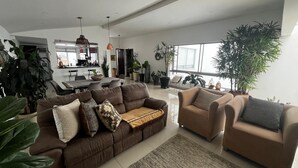 Een ruime woonkamer met een bruine zithoek, een salontafel en een vloerkleed. Er staan diverse potplanten en een groot raam zorgt voor natuurlijk licht.