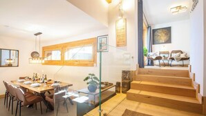 A modern dining area with a wooden table, chairs, and a glass partition. A staircase leads to a living area with a sofa and a coffee table. - Morzna Etoile hosted by More Mountain Morzine | Luxury 3 Bed 3 Bath | Sleeps 6 | Stylish Split level duplex apartment in a newly renovated farmhouse, just 2 mins from Morzine centre. Terrace with views and Outdoor Jacuzzi. (Morzine)