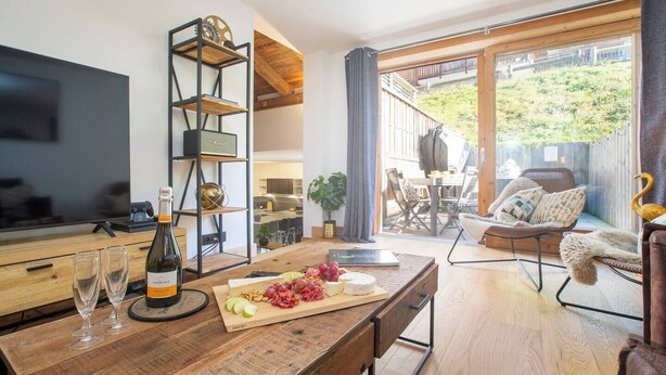 A modern kitchen with a wooden countertop, wine glasses, and a bottle of wine. There is a dining area with a table and chairs, and a view of the outdoors through large windows. - Morzna Etoile hosted by More Mountain Morzine | Luxury 3 Bed 3 Bath | Sleeps 6 | Stylish Split level duplex apartment in a newly renovated farmhouse, just 2 mins from Morzine centre. Terrace with views and Outdoor Jacuzzi. (Morzine)