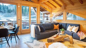 A cozy, well-lit living room with a dark gray sofa, a wooden coffee table, and a dining area with chairs and a table set for a meal.