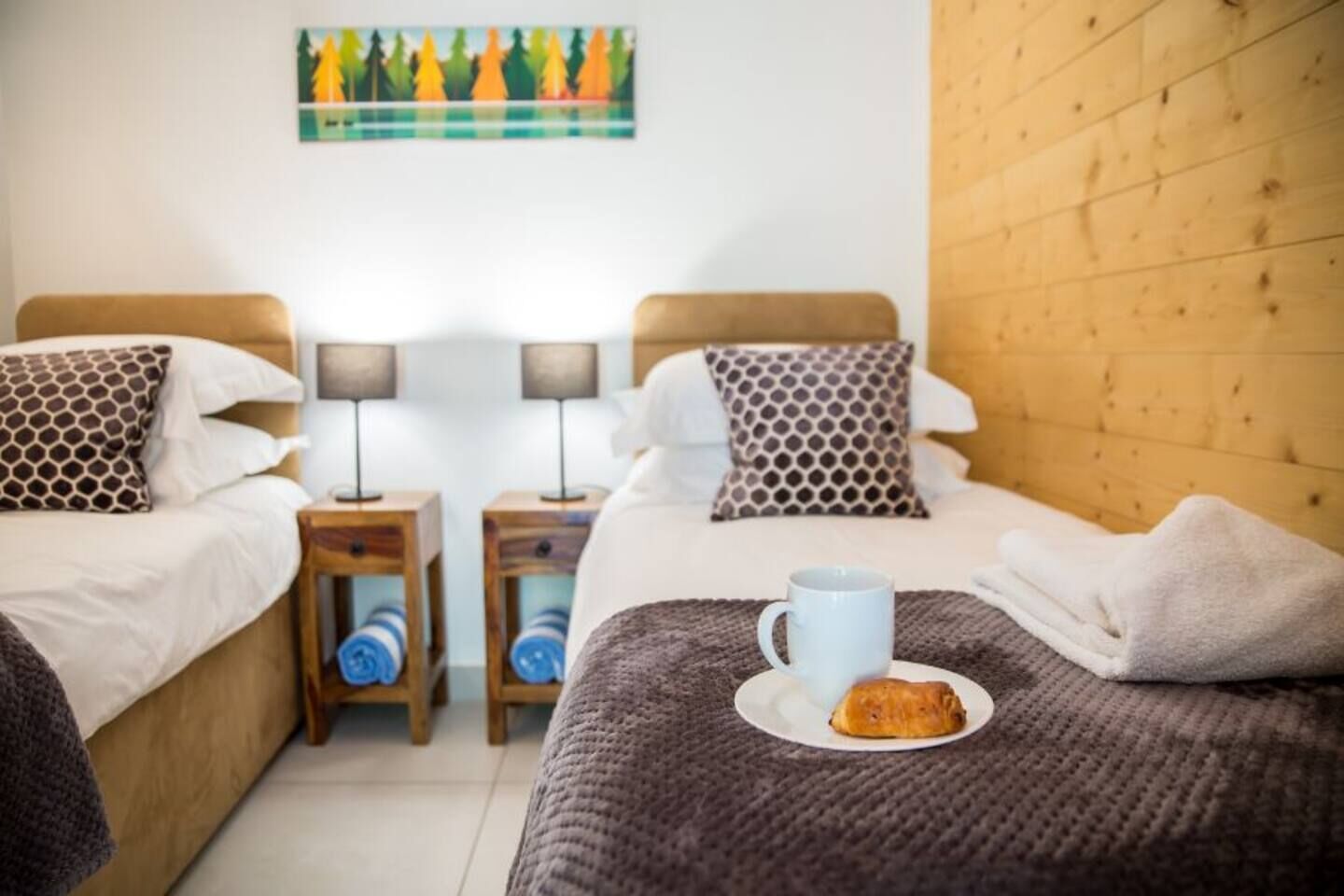 A hotel room with two beds, each with patterned pillows and a bedside table with a lamp. A plate with a pastry and a mug are on the bed.