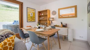 Een eetgedeelte met een houten tafel gedekt voor vier personen, stoelen, een klein keukentje met een koelkast en een groot raam met uitzicht op het omliggende landschap.