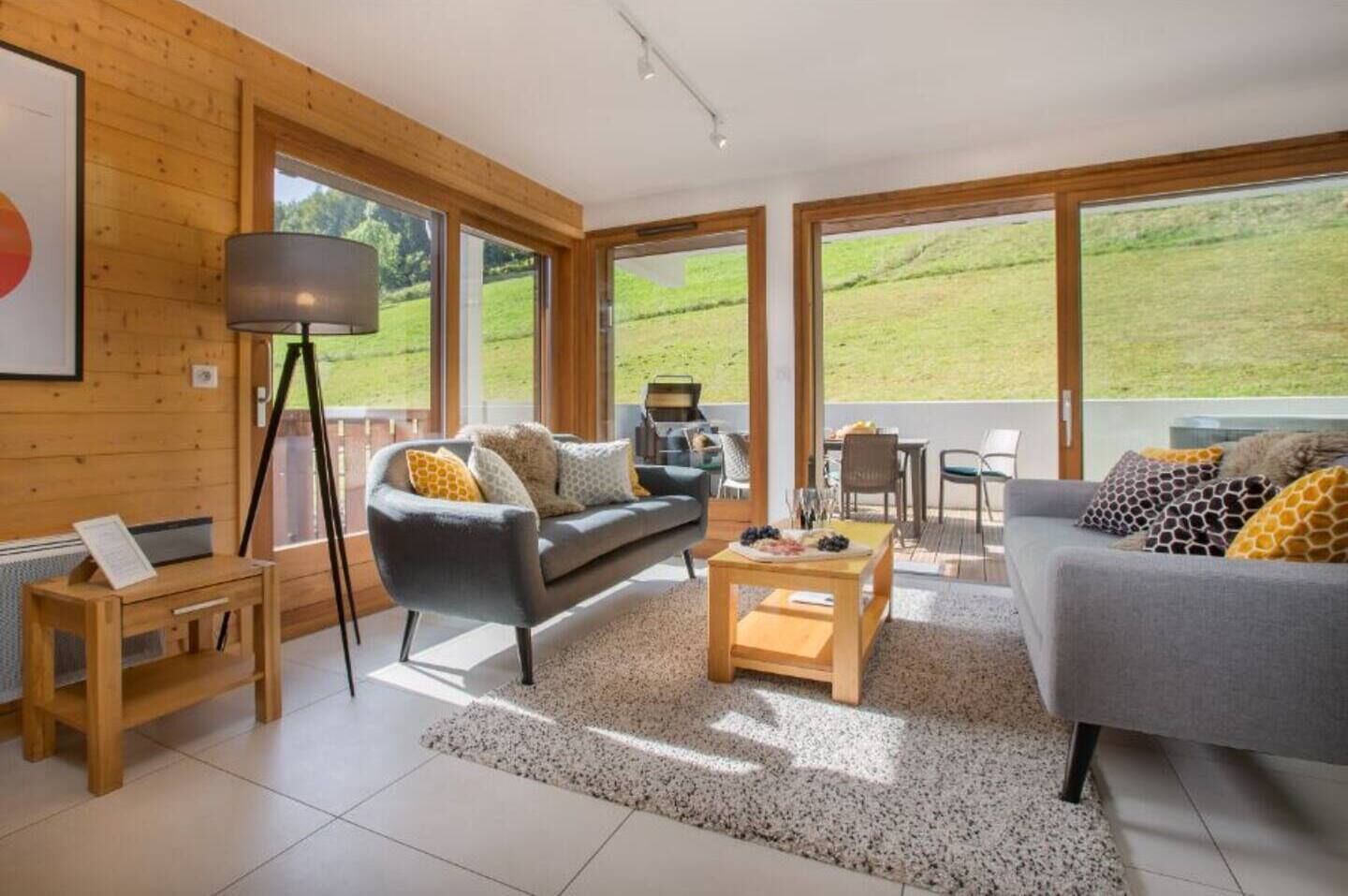 A spacious living room with a grey sofa, a wooden coffee table, and a floor lamp. Large windows offer a view of a green landscape.