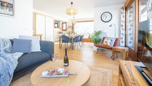 Een moderne woonkamer met een bank, salontafel en televisie. Er is een zithoek met stoelen en een tafel, en een keuken op de achtergrond.