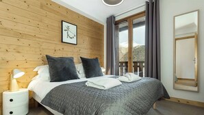 Een slaapkamer met een houten wand, een bed met een grijze quilt, twee kussens, een bedlampje en een raam met gordijnen.