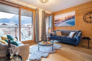 A cozy living room with a blue sofa, a glass coffee table, and a view of snowy mountains through large windows. - Apartment Ressachaux hosted by More Mountain, Morzine | Luxury 3 Bed 3 Bath + Bunk Room | Sleeps 8. Spectacular Apartment in the centre of town opposite the Super Morzine Telecabine. You cannot beat this for size and location! (Morzine)