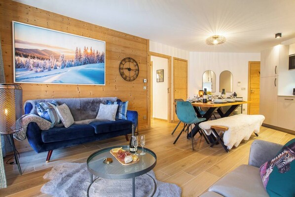 Un salon chaleureux avec un canapé bleu, un coin repas doté d’une table en bois et un grand tableau représentant un paysage enneigé.