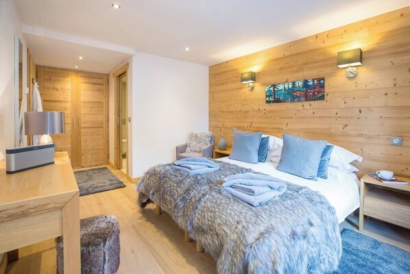 A modern bedroom with a wooden wall panel, a bed with blue bedding, a desk with a lamp, and a painting on the wall.
