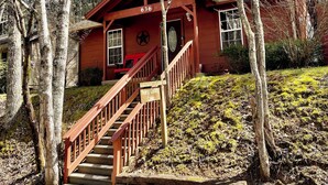 A cabin with a wooden staircase leading to a front door, surrounded by trees and a grassy area.