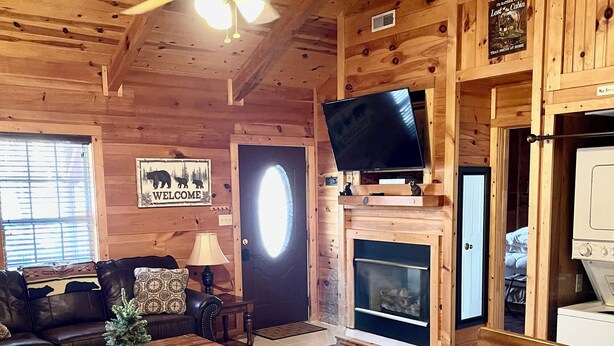 A cozy cabin interior with a wooden ceiling, a fireplace, a television, and a dining area.