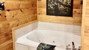 A wooden cabin bathroom with a large bathtub, a framed picture of a bear on a tree, and a towel hanging on the tub.