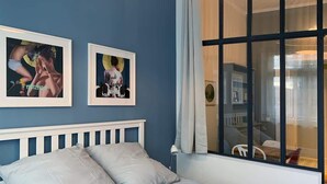 A bedroom with a bed, two framed pictures on the wall, a window with curtains, and a view of a dining area.
