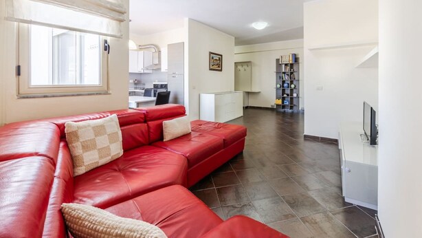 A modern living room with a red leather sofa, a kitchen in the background, and a bookshelf. - Spacious Stay in the Heart of Tirana 2BR City View (Tirana)