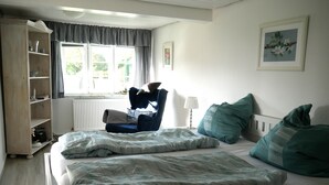 Un dormitorio con cama, una silla, un radiador, una ventana y un cuadro en la pared.