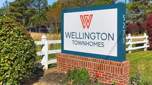 A sign for Wellington Townhomes with a logo, a brick base, and a white picket fence in the background. - Bright 2Bd 1Bt Homes in Central Wellington (College Park)