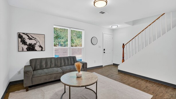 A living room with a gray sofa, a round coffee table with a vase, a window with blinds, a staircase, and a door. - Bright 2Bd 1Bt Homes in Central Wellington (College Park)