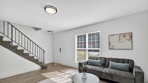 A modern living room with a staircase, a sofa, a coffee table, and a window with blinds. - Bright, Spacious 2Bd 2.5Bt Townhome Interior (College Park)