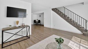 A modern living room with a flat-screen TV, a console table, a staircase, and a dining area in the background. - Bright, Spacious 2Bd 2.5Bt Townhome Interior (College Park)