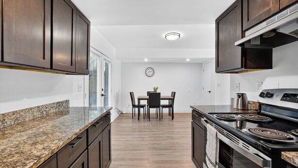 A modern kitchen with dark wooden cabinets, granite countertops, a built-in stove, and a dining area with a table and chairs. - Bright, Spacious 2Bd 2.5Bt Townhome Interior (College Park)