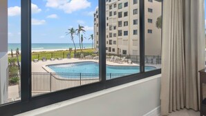 Ein Hotelzimmer mit großem Fenster und Blick auf Pool, Strand und Palmen.