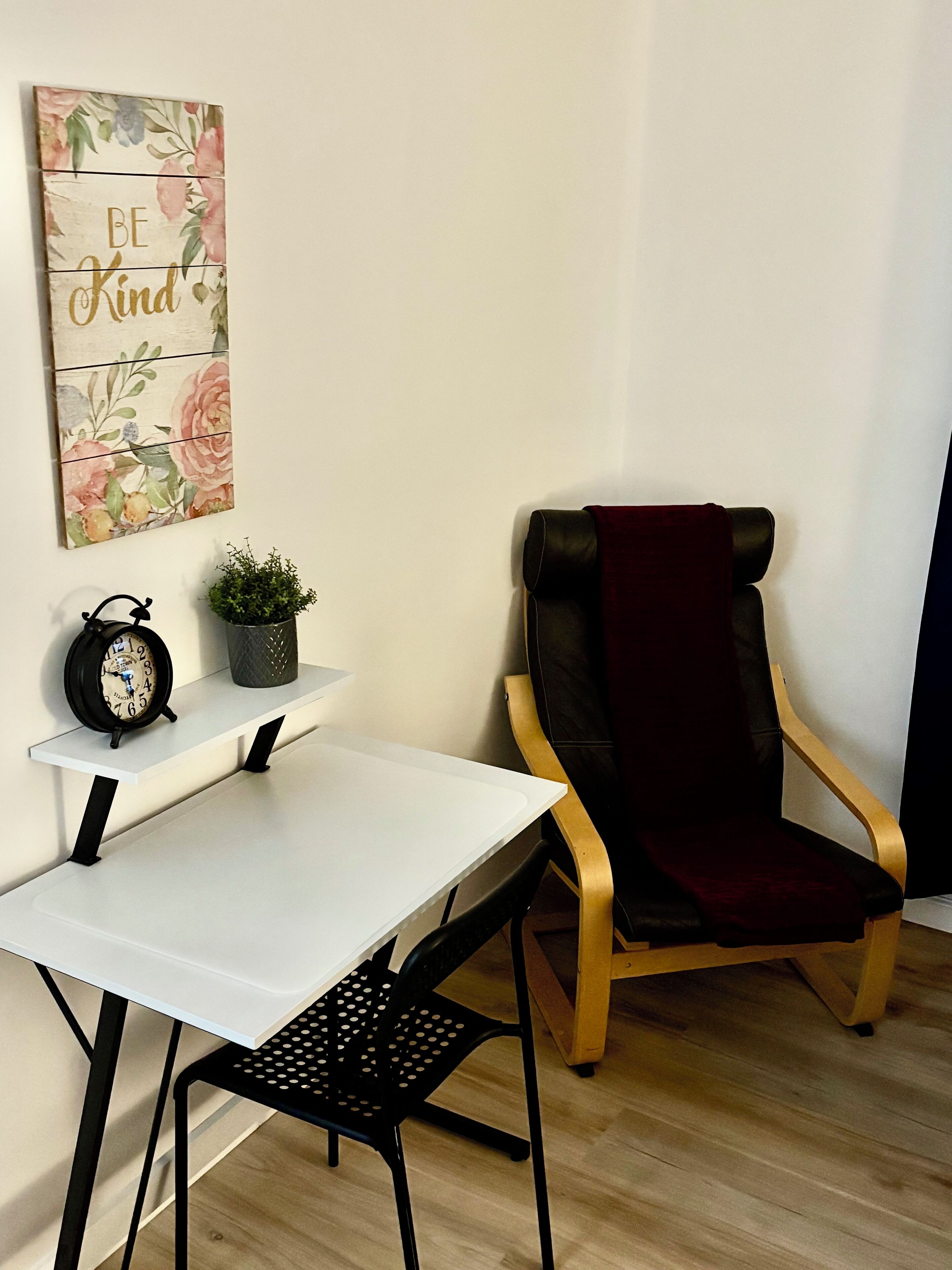 Quarto com uma mesa branca, uma cadeira preta perfurada, uma cadeira vermelho escuro, uma pequena planta em vaso e um relógio fixado na parede.