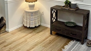 Une pièce avec un mur blanc, un plancher en bois, une lampe de table en dentelle, une console en bois foncé avec un plateau, et une fenêtre à cadre blanc.