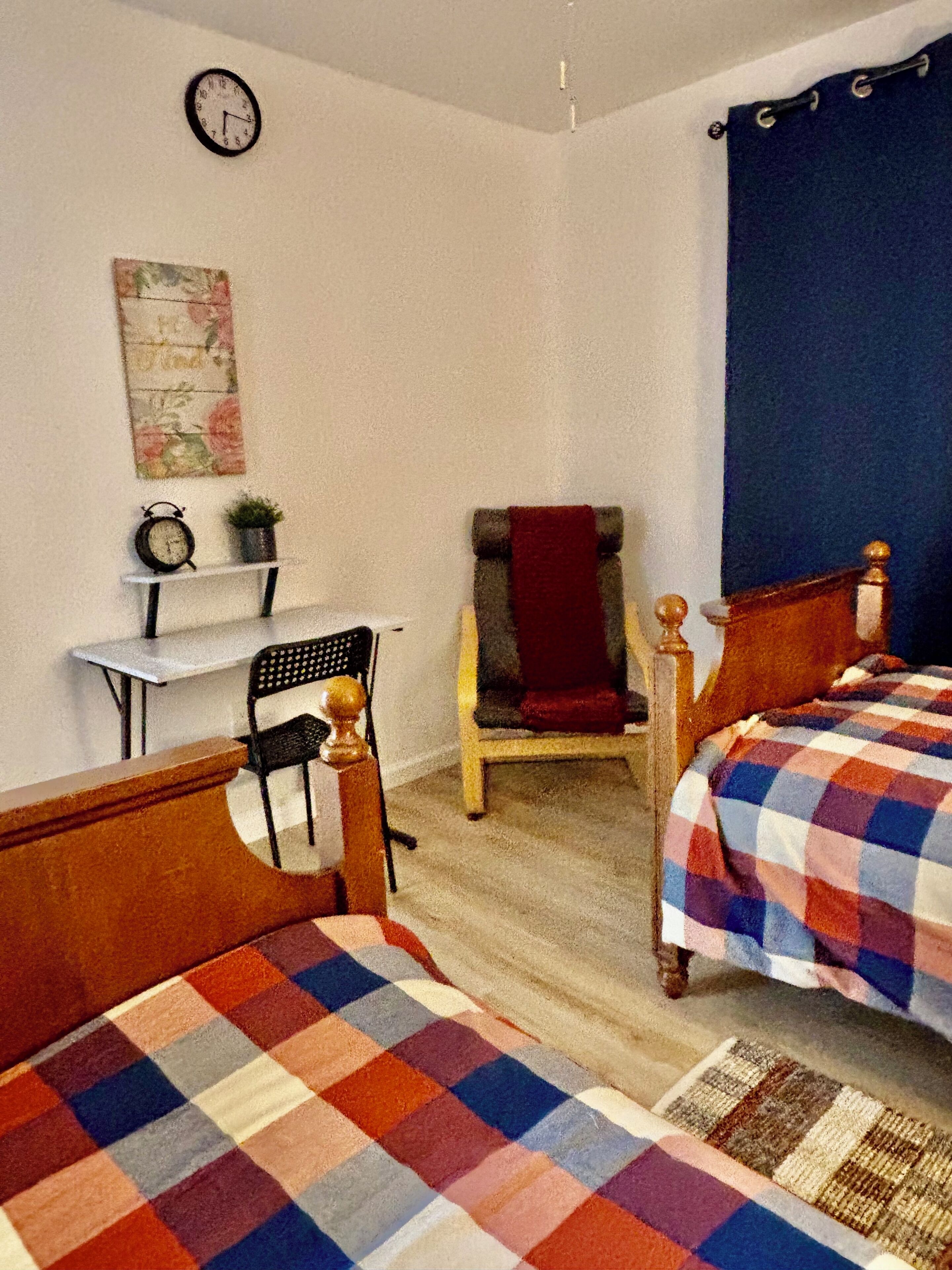 Una habitación con dos camas, un escritorio con una silla, un reloj y un cuadro floral en la pared.