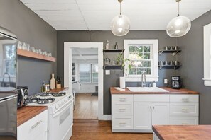 A modern kitchen with white cabinetry, a wooden countertop, and a gas stove. There are hanging pendant lights, a window with a view, and shelves with kitchenware.