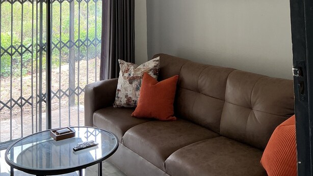 A living room with a brown leather sofa, a glass coffee table, a remote control, and a framed abstract painting on the wall. - Cosy modern cottage only 10 minutes from the city centre (Mutare)