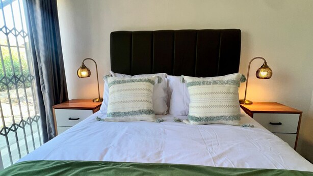 Una cama bien hecha con una colcha verde, dos lámparas de noche y una ventana con cortinas.